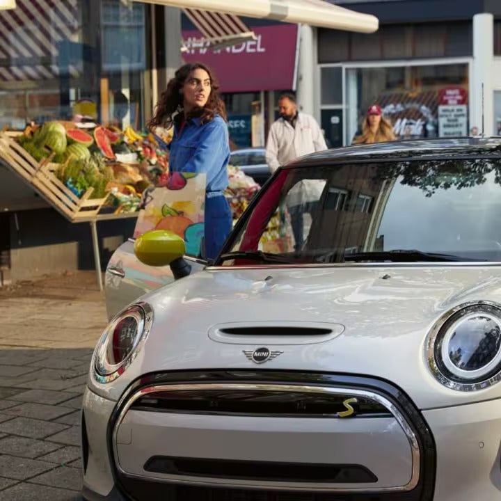 Three-quarters front view of a MINI Electric vehicle parked on a brick surface with a fruit stand and retail storefronts in the background along with people standing nearby.
