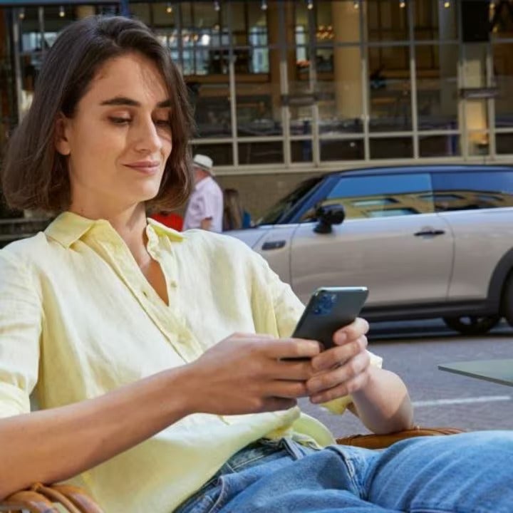 Woman in yellow button-down shirt and jeans sitting down while holding a smartphone and glancing at it, with a grey MINI vehicle parked on a street surface in the background.