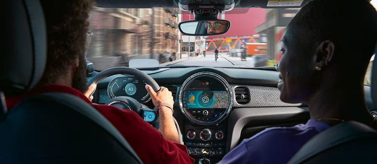 View of two men glaring onwards through the windshield of a MINI vehicle while on the road in an urban setting, pictured from the back row of a MINI vehicle.